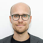 The image shows a light-skinned man with glasses, a short beard, and a bald head. He is wearing a dark T-shirt and a gray jacket, giving off a neat and professional appearance. His expression is calm and friendly.