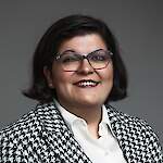 A smiling woman with short dark hair, wearing glasses, a white collared shirt, and a patterned jacket, photographed against a neutral background.