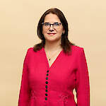 A woman with shoulder-length brown hair and patterned round glasses smiles softly while wearing a bright pink textured top against a plain beige background.