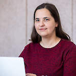 A woman with straight, shoulder-length brown hair sitting at a table in front of a laptop.
