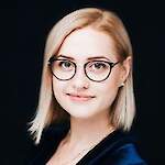 A professional portrait of a blonde woman with round black glasses, wearing a deep blue velvet top and a subtle necklace against a dark background.